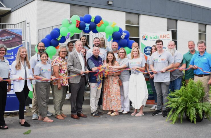 A group of people stand in front of a building holding a ribbon for a ribbon-cutting ceremony with balloons overhead.
