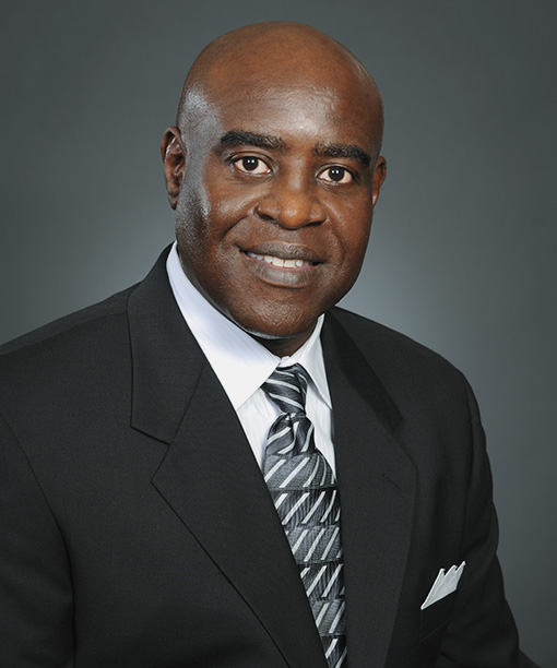 A man in a dark suit and patterned tie poses for a professional headshot against a gray background.