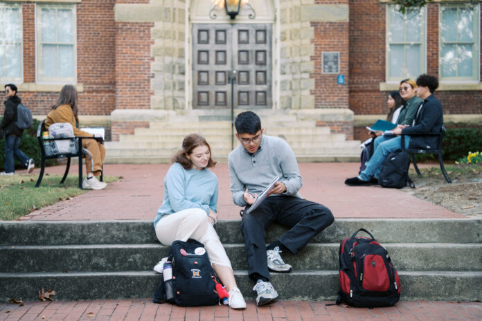 Two Mercer students sit on steps outside a building, studying together with backpacks nearby; others sit on benches in the background.