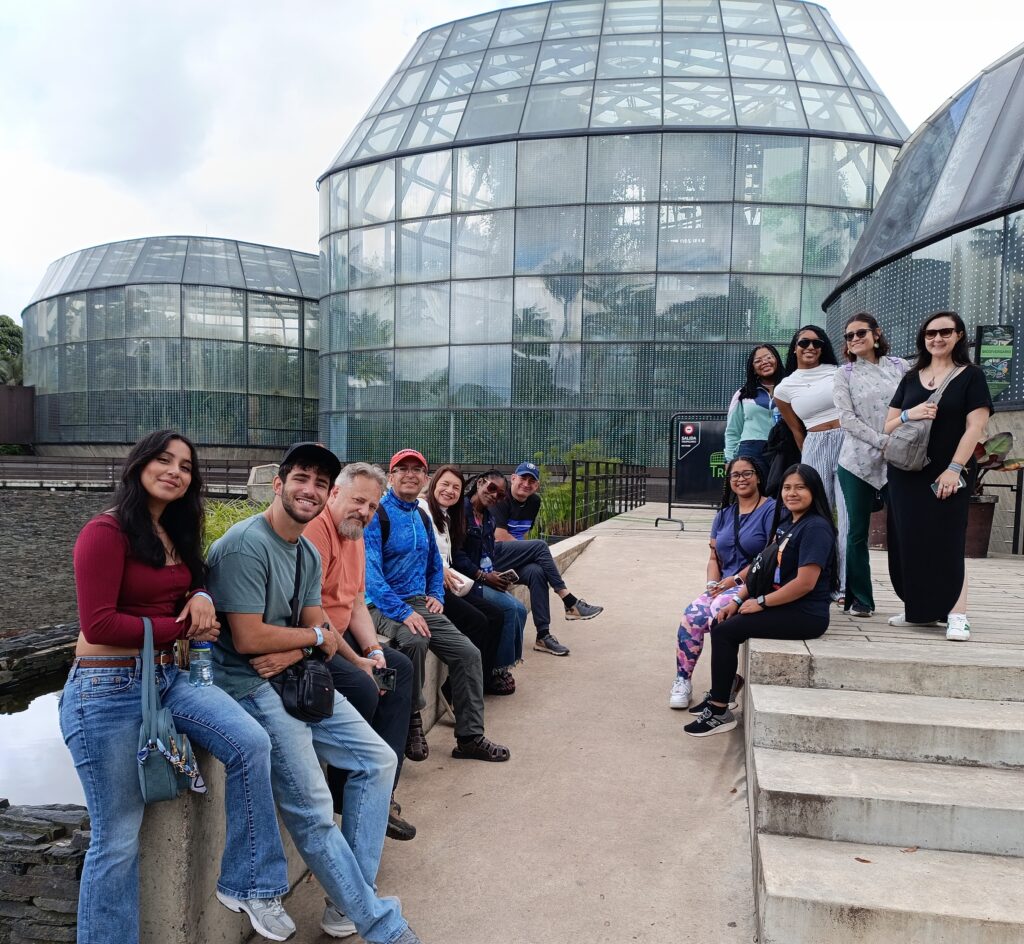 A group of people sits and stands outside glass greenhouse domes, posing for a group photo on a cloudy day.