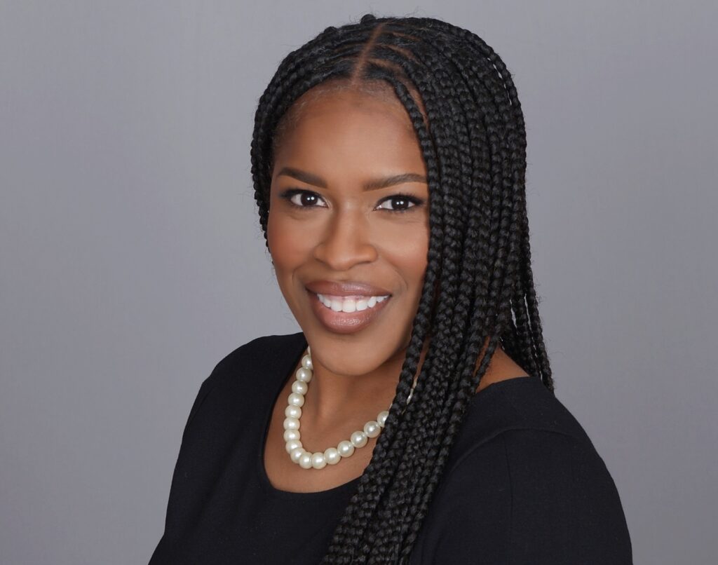 Woman with long braided hair wearing a black dress and pearl necklace, smiling against a plain gray background.