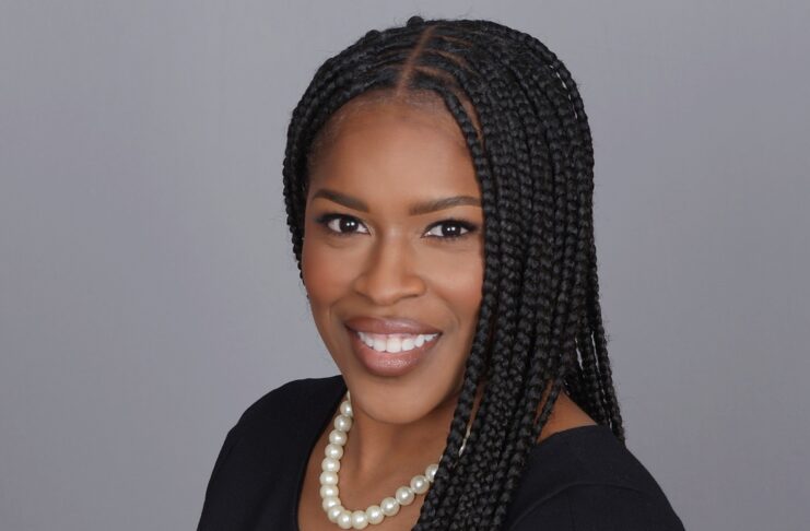 Woman with long braided hair wearing a black dress and pearl necklace, smiling against a plain gray background.