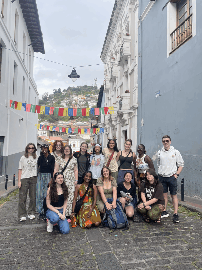 A group of people pose for a photo on a cobblestone street with colorful flags hanging above and a hillside in the background.
