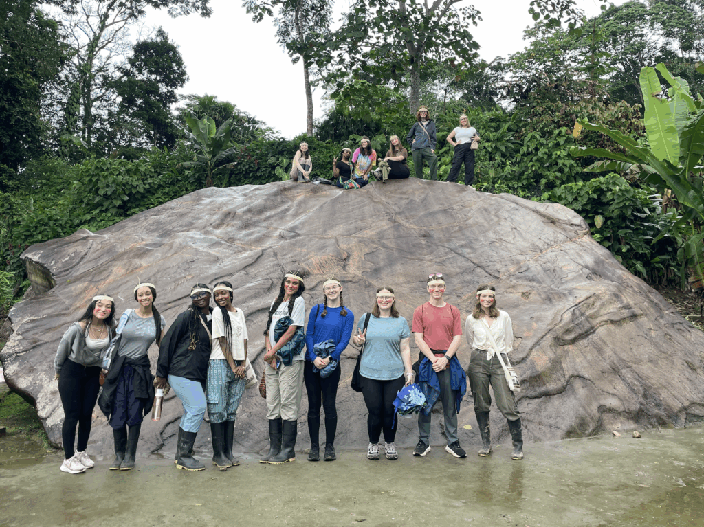 A group of people pose in front of and on top of a large rock in a lush, green outdoor setting.