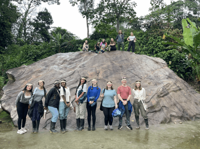 A group of people pose in front of and on top of a large rock in a lush, green outdoor setting.