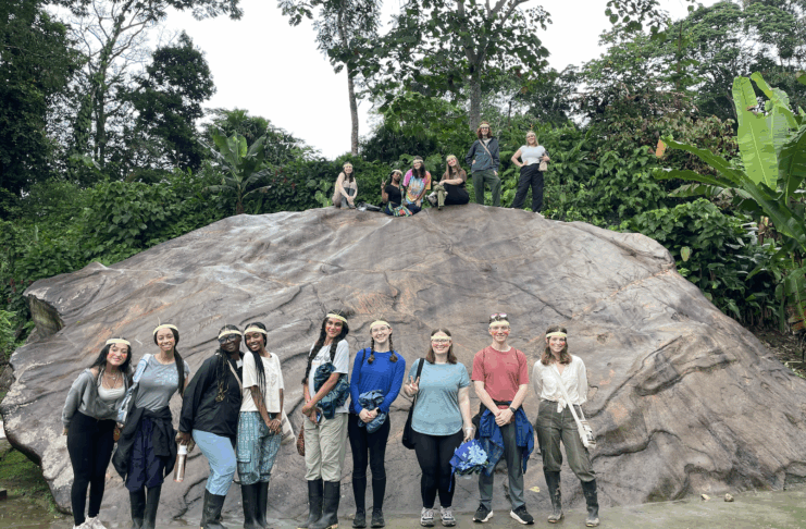 A group of people pose in front of and on top of a large rock in a lush, green outdoor setting.
