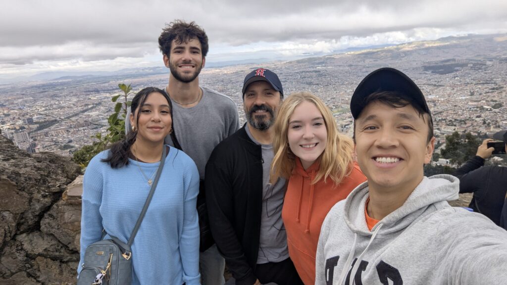 Five people pose for a group selfie with a cityscape and cloudy sky in the background.