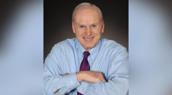 Dr. Richard Swindle strengthened University through his leadership | Mercer Legends Older man in blue dress shirt and purple tie, smiling, sits with arms folded on a table against a dark background.