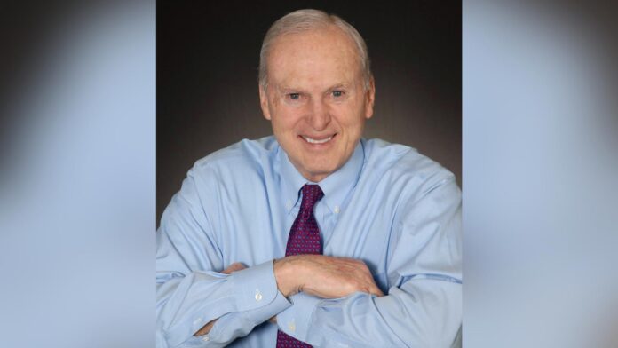 Older man in blue dress shirt and purple tie, smiling, sits with arms folded on a table against a dark background.