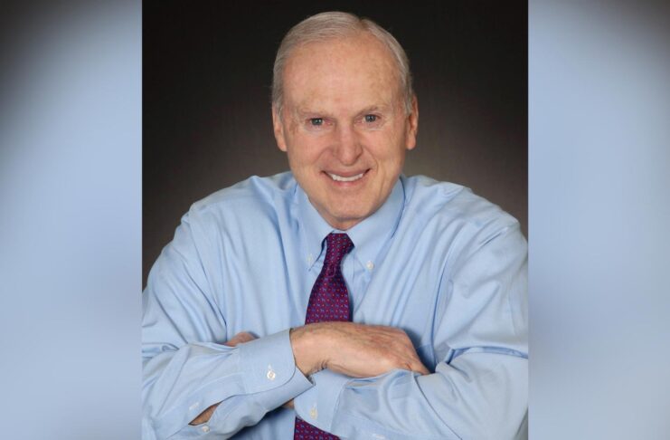 Older man in blue dress shirt and purple tie, smiling, sits with arms folded on a table against a dark background.