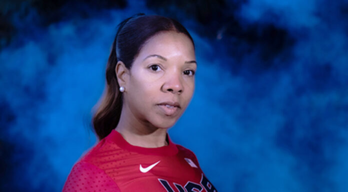 Mercer law alumna balances legal career with world-class hurdles A woman in a red long-sleeve USA athletic shirt stands in front of a blue, smoky background, looking at the camera.