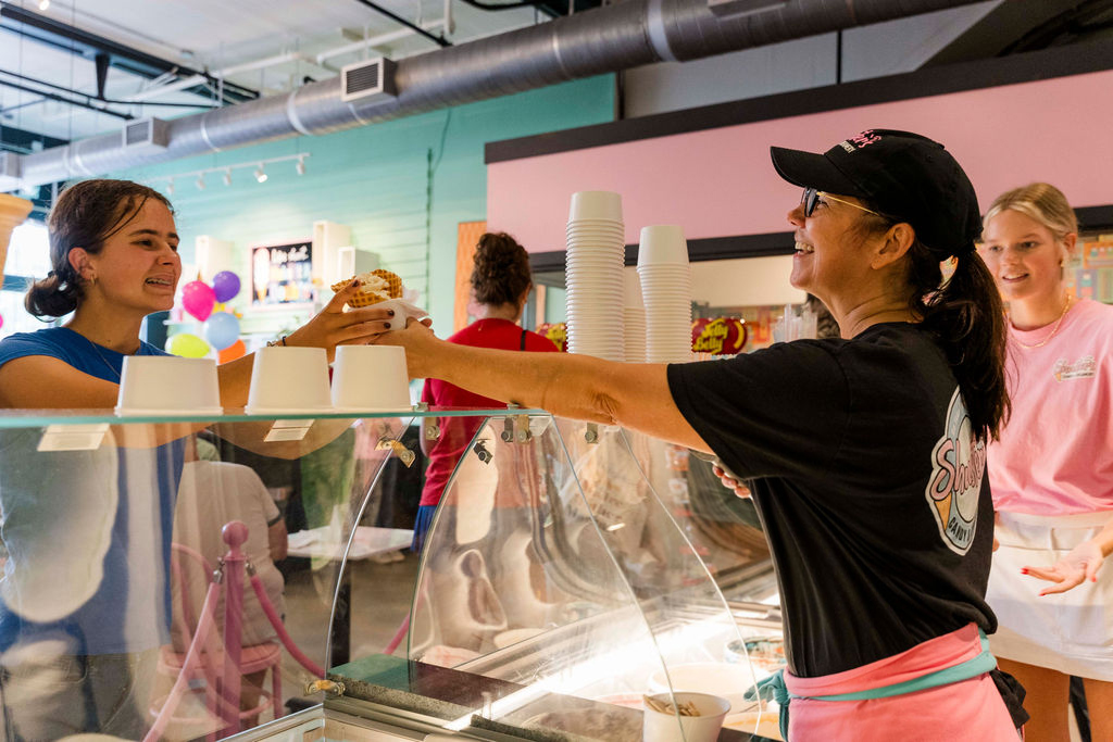 A worker hands an ice cream sundae to a smiling customer across the counter in an ice cream shop.