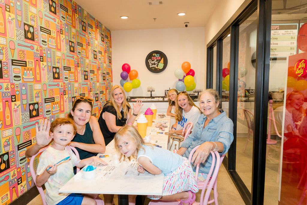 A group of people, including children and adults, sit at a table in an ice cream shop decorated with colorful balloons and wallpaper.