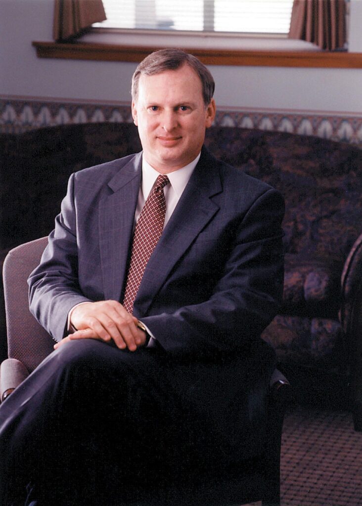 A man in a dark suit sits in an office chair with his legs crossed, posing for a formal portrait.