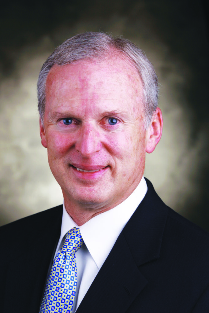 A middle-aged man in a suit and tie poses for a professional headshot against a blurred background.