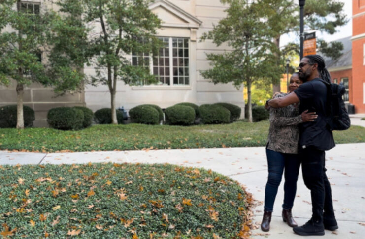 Two people stand on a sidewalk outside a building, hugging each other amid greenery and fallen leaves.