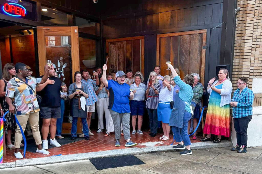 A group of people stand and cheer outside a storefront with open doors on a damp sidewalk.
