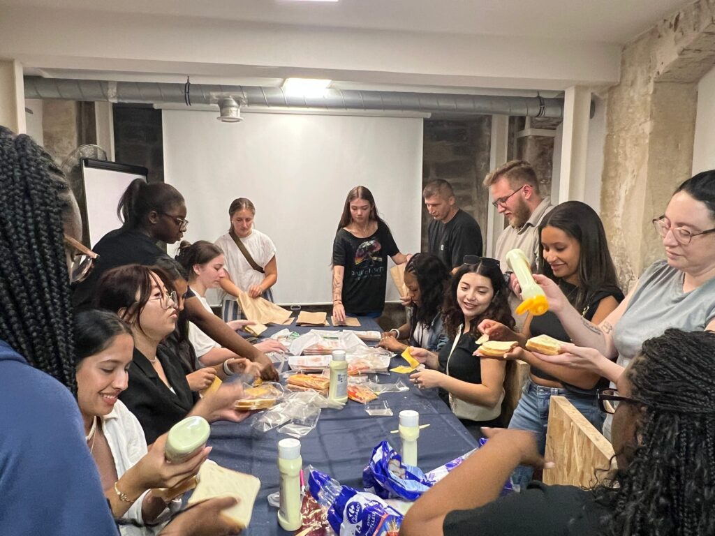 A group of people prepares sandwiches together around a table covered with bread, cheese, and condiments.