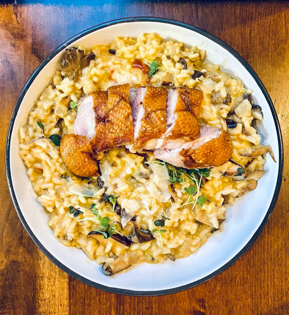 A bowl of mushroom risotto topped with sliced roasted duck breast, garnished with microgreens.