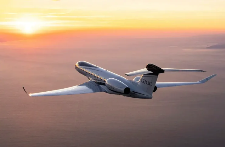 Private jet labeled G700 flying over the ocean at sunset with orange and yellow sky in the background.
