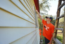 Annual ‘Be a Good NeighBear’ Service Day Saturday Person in an orange shirt paints the exterior siding of a house with a roller brush during daytime.