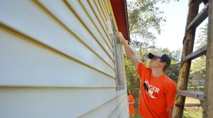 Annual ‘Be a Good NeighBear’ Service Day Saturday Person in an orange shirt paints the exterior siding of a house with a roller brush during daytime.