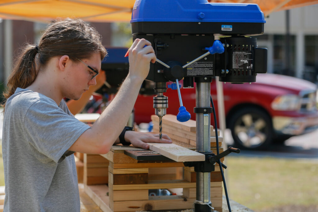 Mercer student uses a drill press to make holes in a wooden board outdoors.