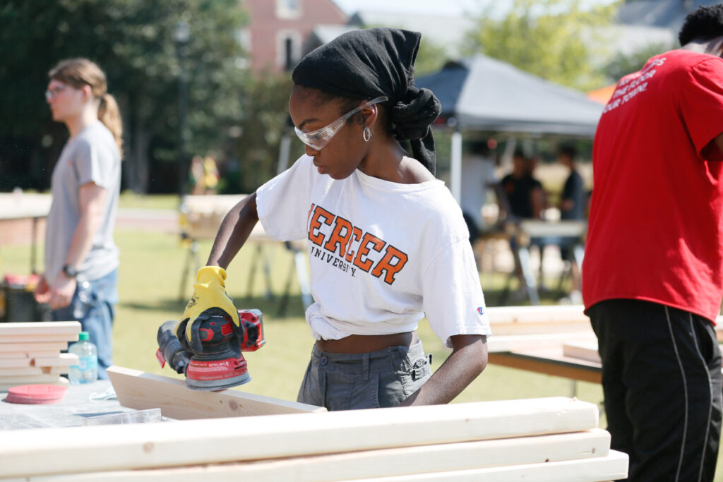Mercer student wearing safety goggles sands a wooden board with a power tool outdoors.