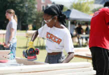 10 community service projects that helped define MerServe Mercer student wearing safety goggles sands a wooden board with a power tool outdoors.