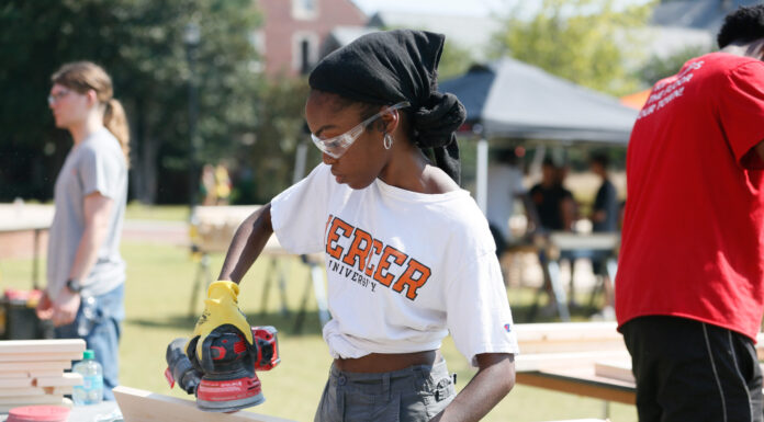 10 community service projects that helped define MerServe Mercer student wearing safety goggles sands a wooden board with a power tool outdoors.