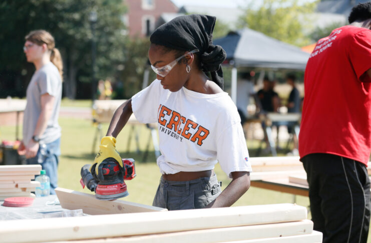 Mercer student wearing safety goggles sands a wooden board with a power tool outdoors.