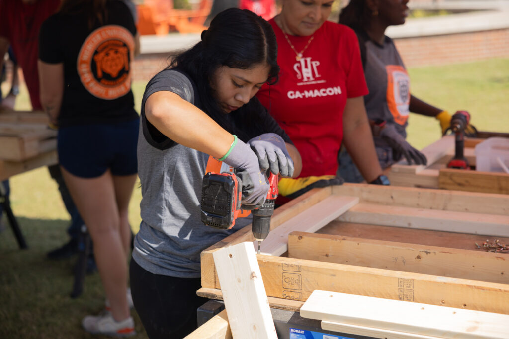 A Mercer student uses a power drill to assemble a wooden frame outdoors, while others work nearby at similar stations.