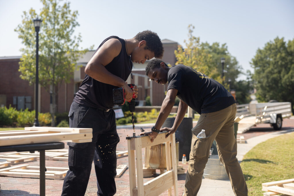 Two Mercer students work together outdoors assembling a wooden structure with power tools on a sunny day.
