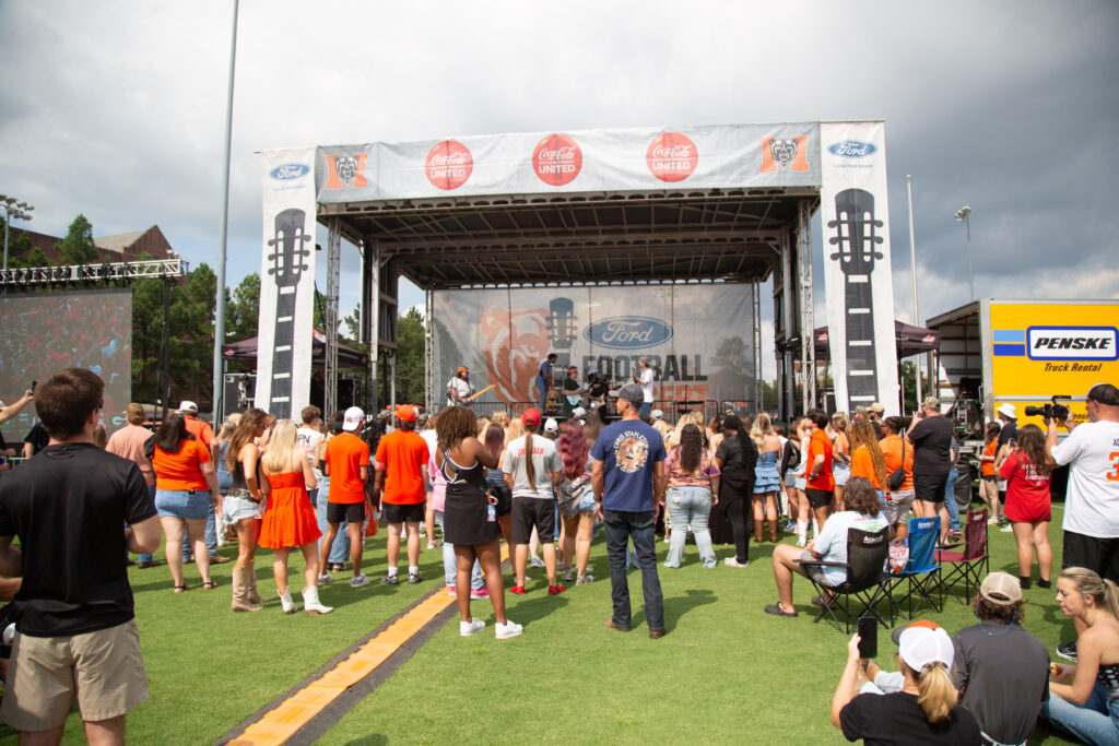 A crowd gathers on a grassy field in front of an outdoor stage during a daytime event with cloudy skies.