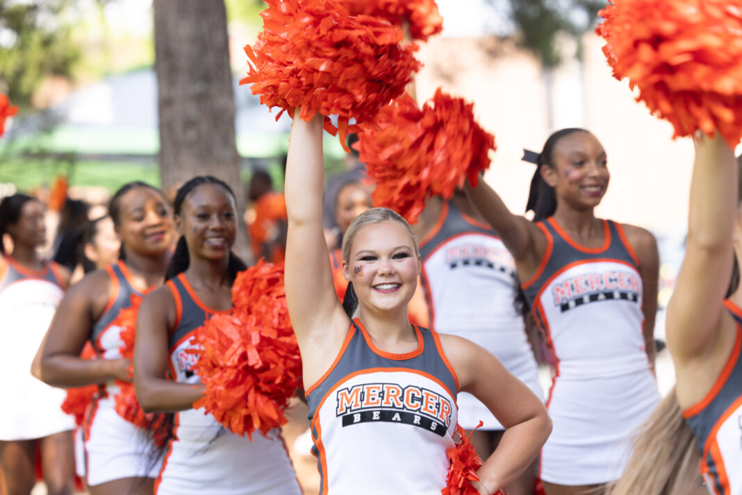 A group of Mercer University cheerleaders in uniform hold orange pom-poms outdoors, one cheering with arm raised.