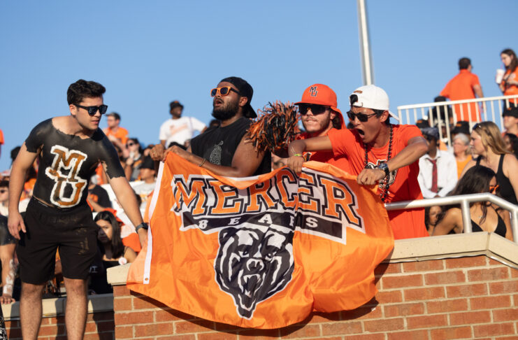 Four fans cheer at a sports event, holding a Mercer Bears flag and wearing Mercer University apparel.
