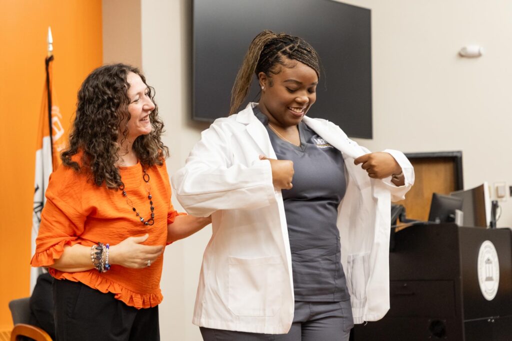 A woman helps a student put on a white coat in a classroom during a ceremony.