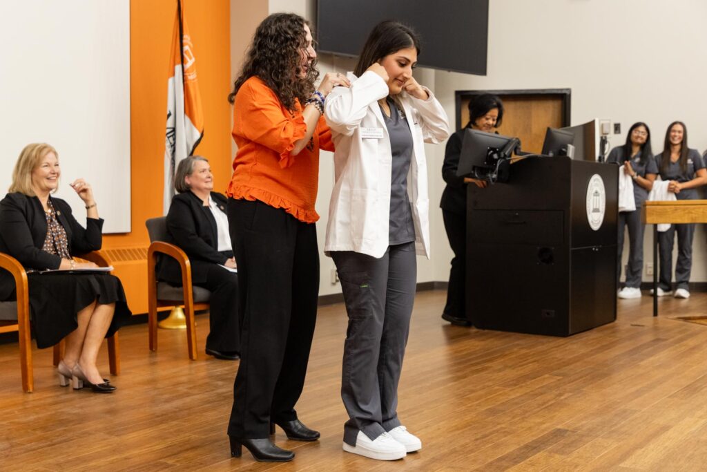A woman helps a student put on a white coat during a ceremony, while others watch and sit in the background.