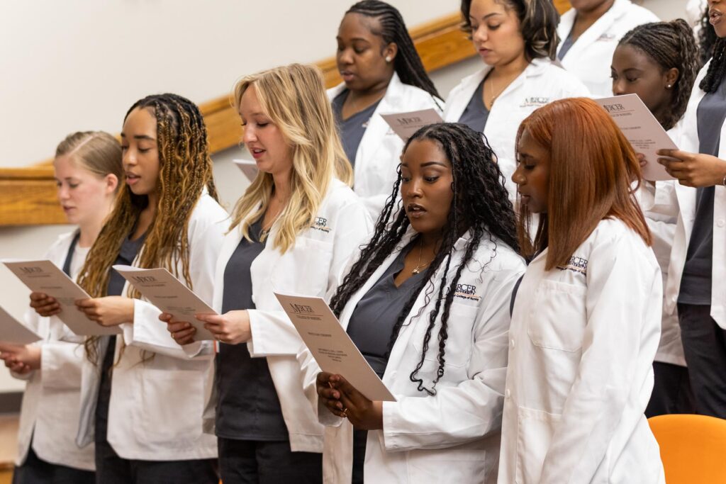 A group of students in white coats stand and read from papers during a ceremony or event in an auditorium.