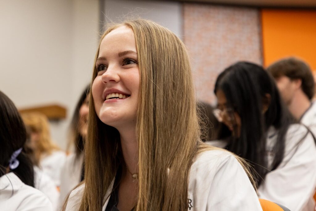 A young woman in a white coat smiles while sitting among peers in a classroom or lecture setting.