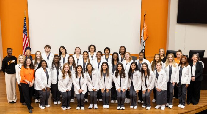 Mercer expands nursing program to Macon campus A large group of students in white coats pose and smile together in front of American and state flags indoors.