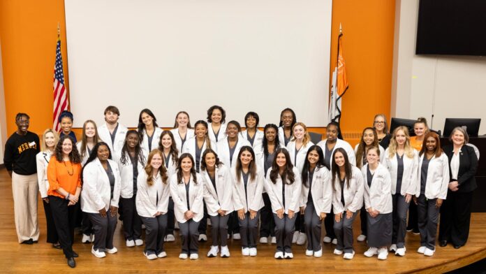 A large group of students in white coats pose and smile together in front of American and state flags indoors.