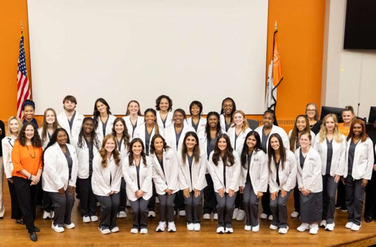 A large group of students in white coats pose and smile together in front of American and state flags indoors.