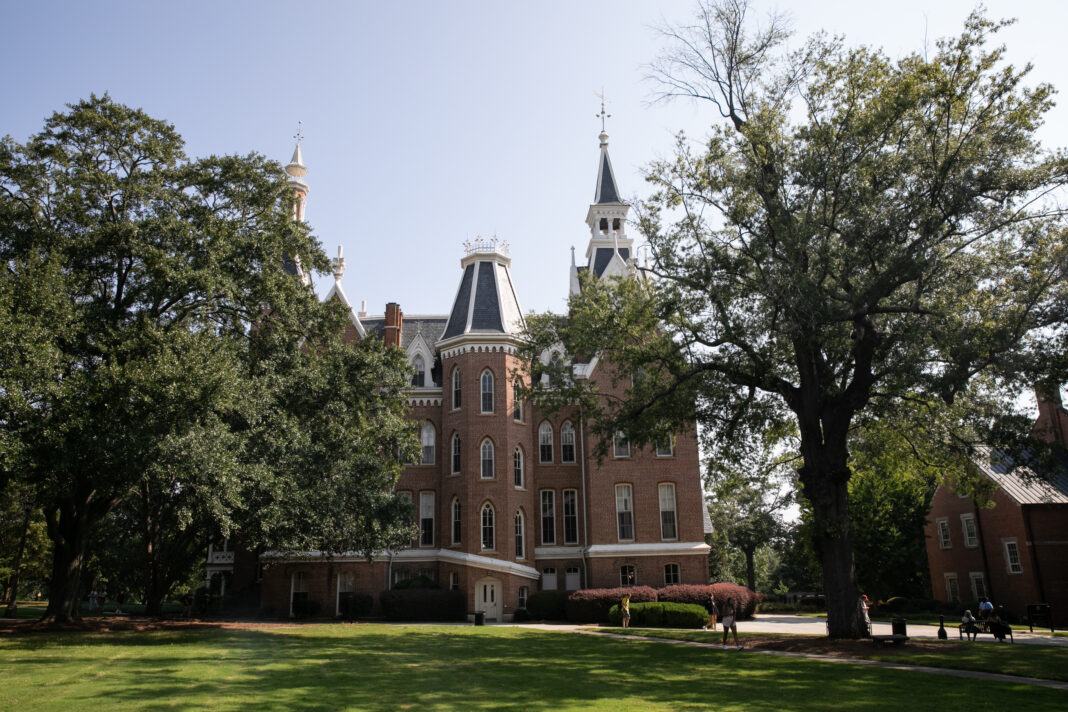 A historic brick building with tall spires stands behind large trees on a sunny day, with people walking nearby.