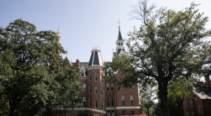 Faculty and Staff Notables | October 2025 A historic brick building with tall spires stands behind large trees on a sunny day, with people walking nearby.
