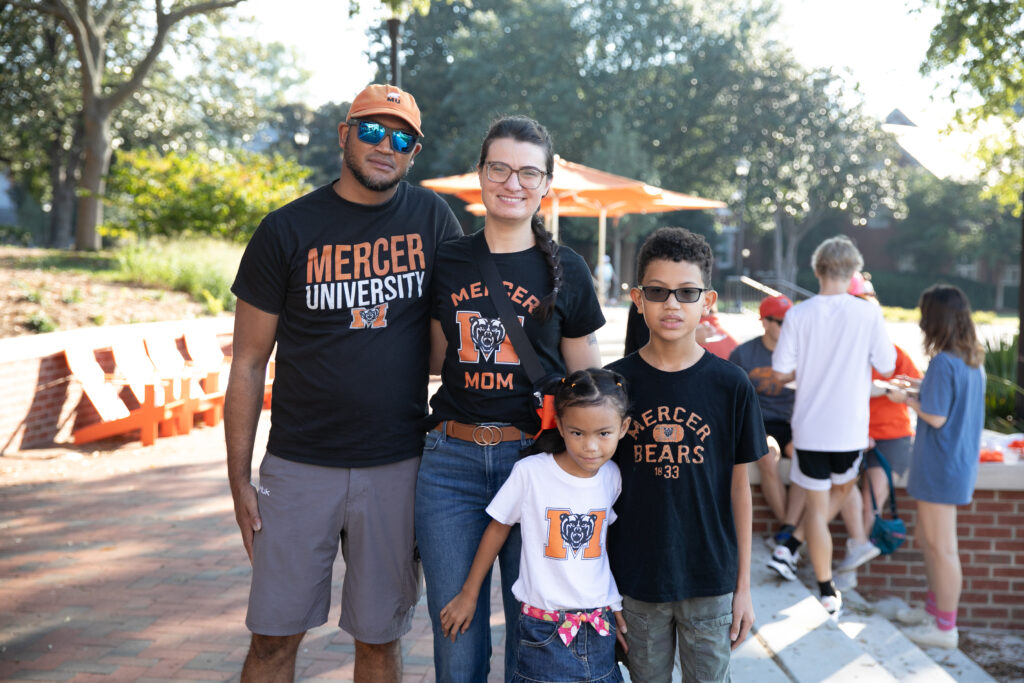 A family of four wearing Mercer University apparel poses outdoors on campus during a sunny day.
