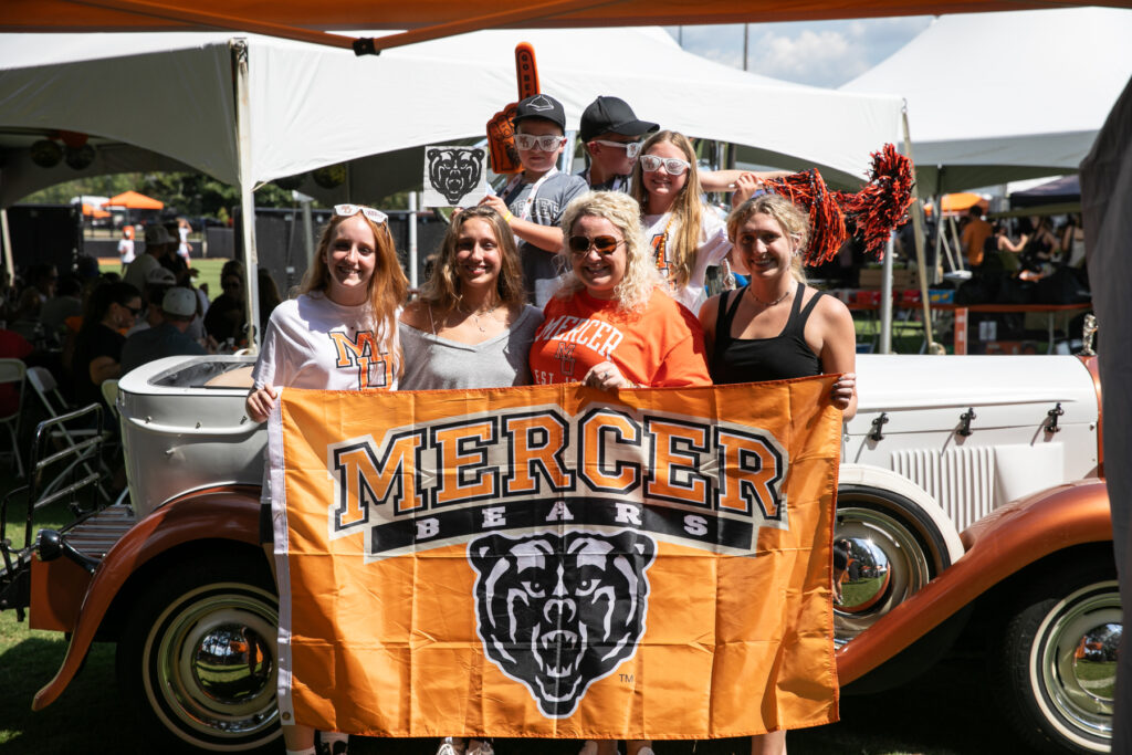 A group of people pose in front of a vintage car, holding a Mercer Bears banner at an outdoor event.