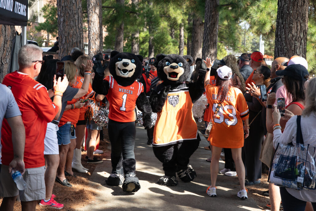 Two bear mascots in orange jerseys walk through a crowd of people wearing orange shirts outdoors among trees.