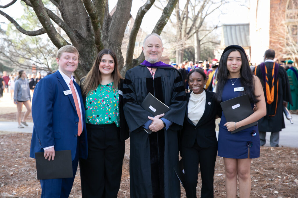 Five people, including one in academic regalia, pose outdoors at a graduation event, holding folders and smiling.