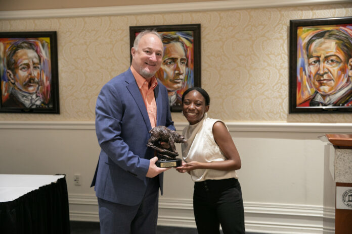 2502_FoundersDay_CiSmith-571 Two people stand indoors holding a trophy together, with framed portraits visible on the wall behind them.